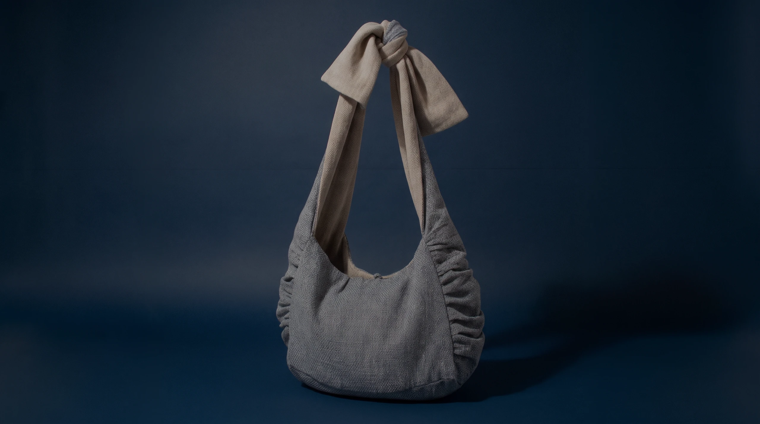 El bolso de hombro gris de asa anudada, sobre un fondo azul oscuro.