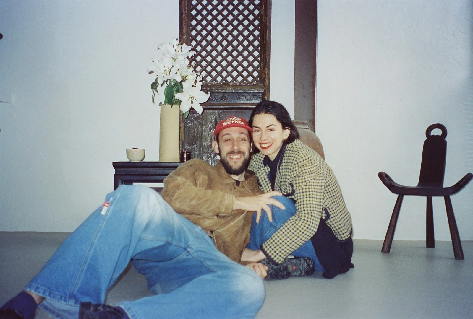 Adriana y Gonzalo sentados en el suelo de una sala blanca, junto a un jarrón con lirios y un taburete de madera.
