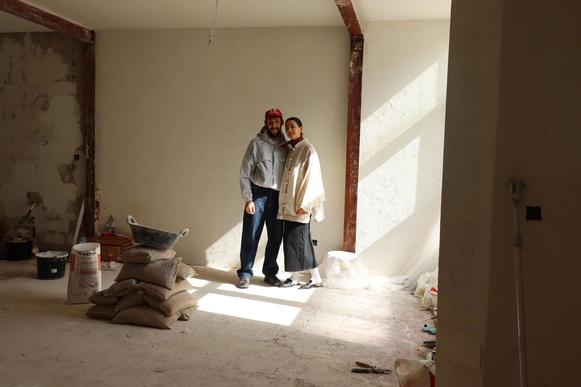 Adriana y Gonzalo de pie en una sala en obras, con sacos y herramientas en el suelo.